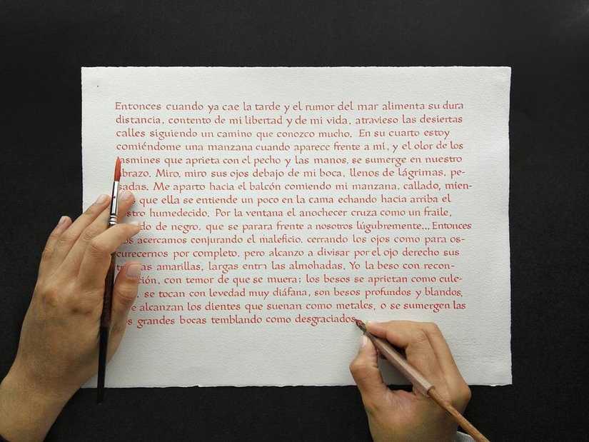 A sheet of paper with many small lines of calligraphy and a hand holding a broad nib pen near the bottom