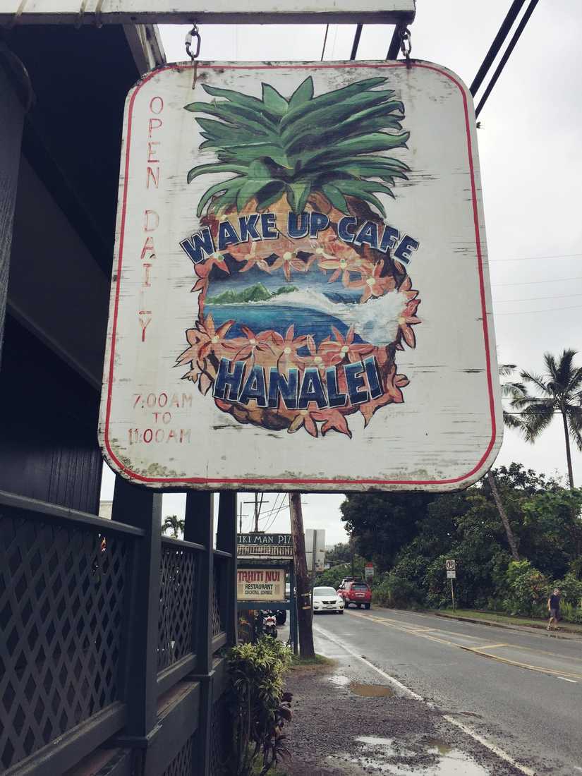 A hanging sign with a detailed painting of a pineapple with a ocean and landscape scene in the centre. The words "Wake up Cafe Hanalei" are incorporated in the illustration.