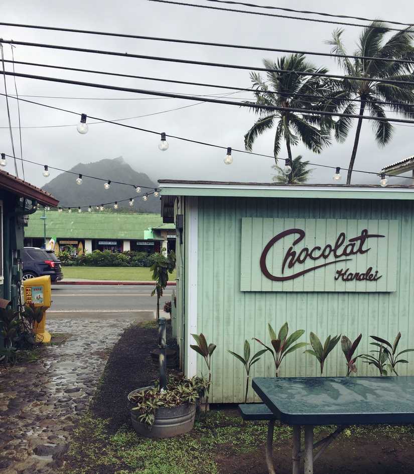 A small building and courtyard with string lights above. The sign is cut from wood and reads "Chocolate Hanalei"